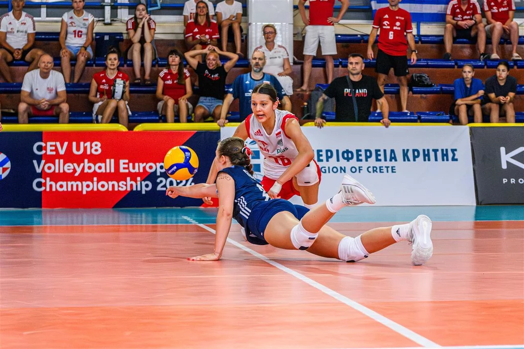 Dwójka zawodniczek na boisku siatkarskim w trakcie dynamicznej akcji, jedna z nich w ślizgu próbuje odbić piłkę, druga zawodniczka z drużyny wspiera akcję; w tle widownia oraz banery turniejowe CEV U18.
