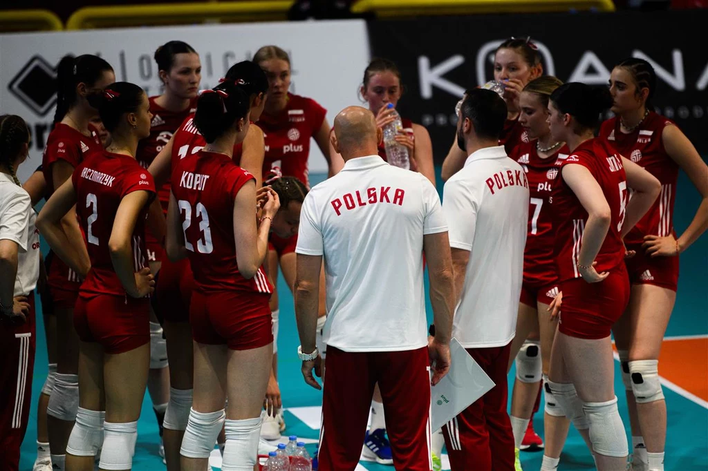 Polska drużyna siatkarska kobiet podczas przerwy w meczu, zawodniczki w czerwonych strojach otaczają trenerów uważnie słuchając wskazówek.