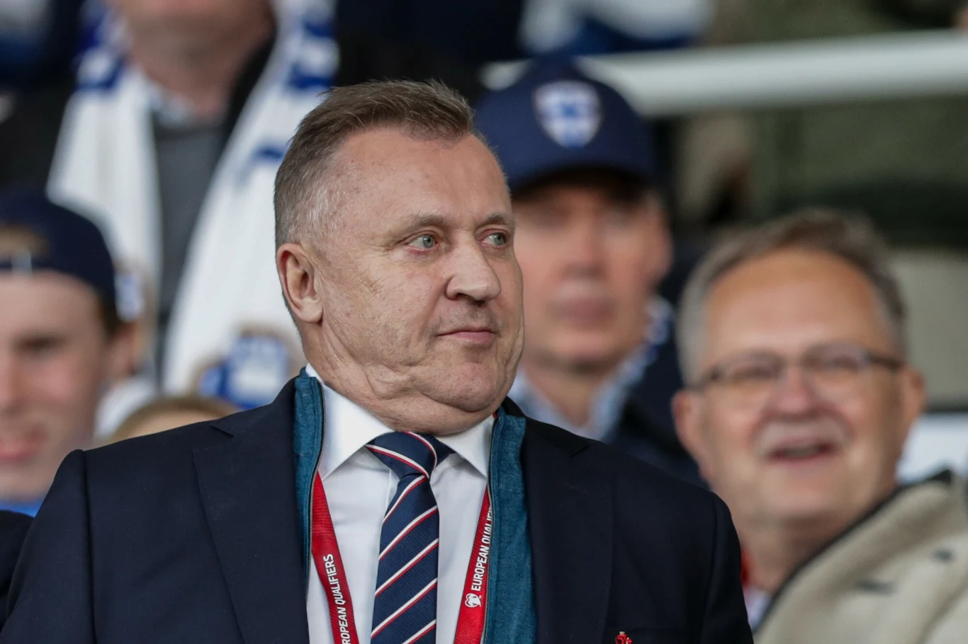 Cezary Kuelsza Mężczyzna w granatowym garniturze, z niebiesko-czerwonym krawatem i szalikiem, stoi na trybunach stadionu, w tle rozmyci kibice uśmiechający się i ubrani w ubrania klubowe.
