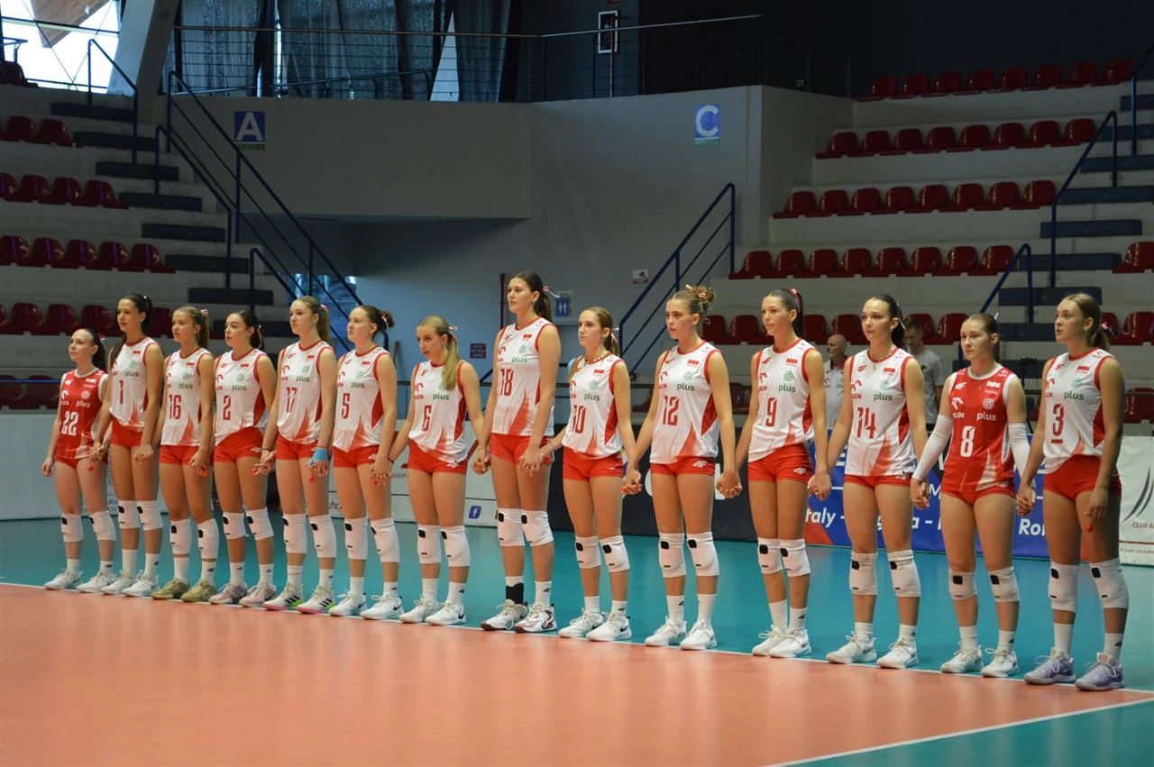 Drużyna siatkarek w czerwono-białych strojach sportowych stoi w równym szeregu na boisku do siatkówki w hali sportowej, sala z pustymi trybunami.