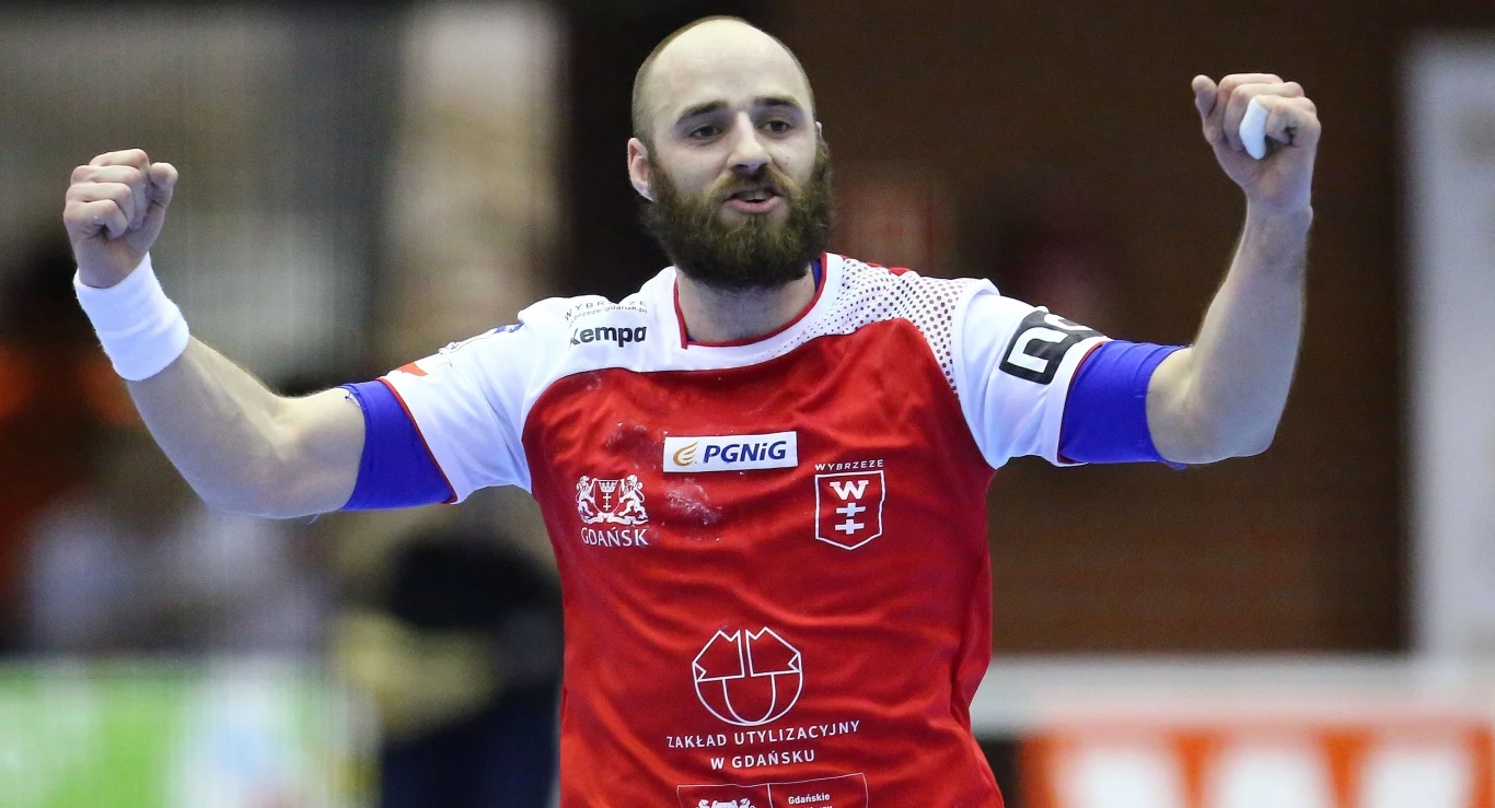 Paweł Niewrzawa Radość zawodnika piłki ręcznej w czerwono-białym stroju sportowym, uniesione ręce w geście triumfu, na tle rozmytego wnętrza hali sportowej.
