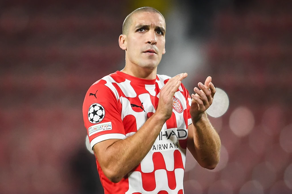 Oriol Romeu Piłkarz w biało-czerwonej koszulce z szachowniczym wzorem, z logo Ligi Mistrzów UEFA na rękawie, klaszczący w dłonie, na tle rozmytego stadionu.