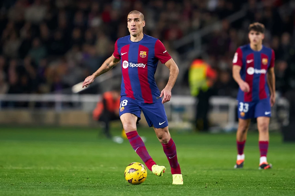Oriol Romeu w barwach FC Barcelona Piłkarz w niebiesko-czerwonym stroju z numerem 18 prowadzi piłkę na boisku, w tle widać innego zawodnika tej samej drużyny oraz kibiców na trybunach.