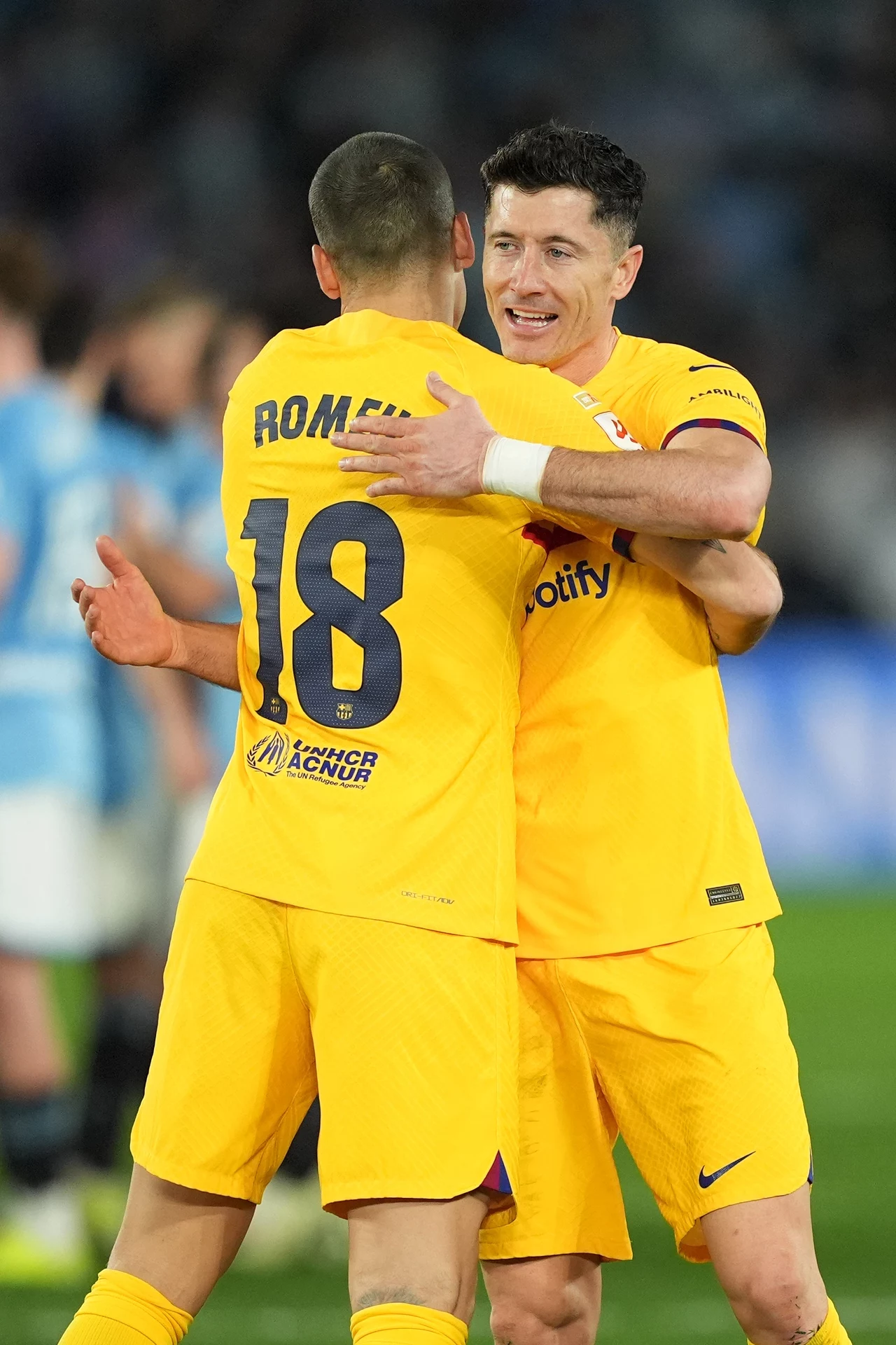 Oriol Romeu i Robert Lewandowski Dwóch piłkarzy w żółtych strojach drużyny podczas przyjacielskiego uścisku na boisku piłkarskim, w tle rozmazane sylwetki innych zawodników i kibiców