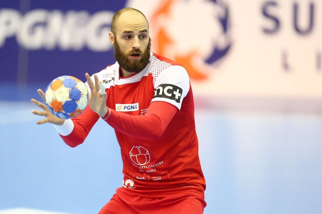 Paweł Niewrzawa Zawodnik piłki ręcznej w czerwonym stroju z piłką w dłoniach, koncentrujący się na grze podczas meczu na hali sportowej.