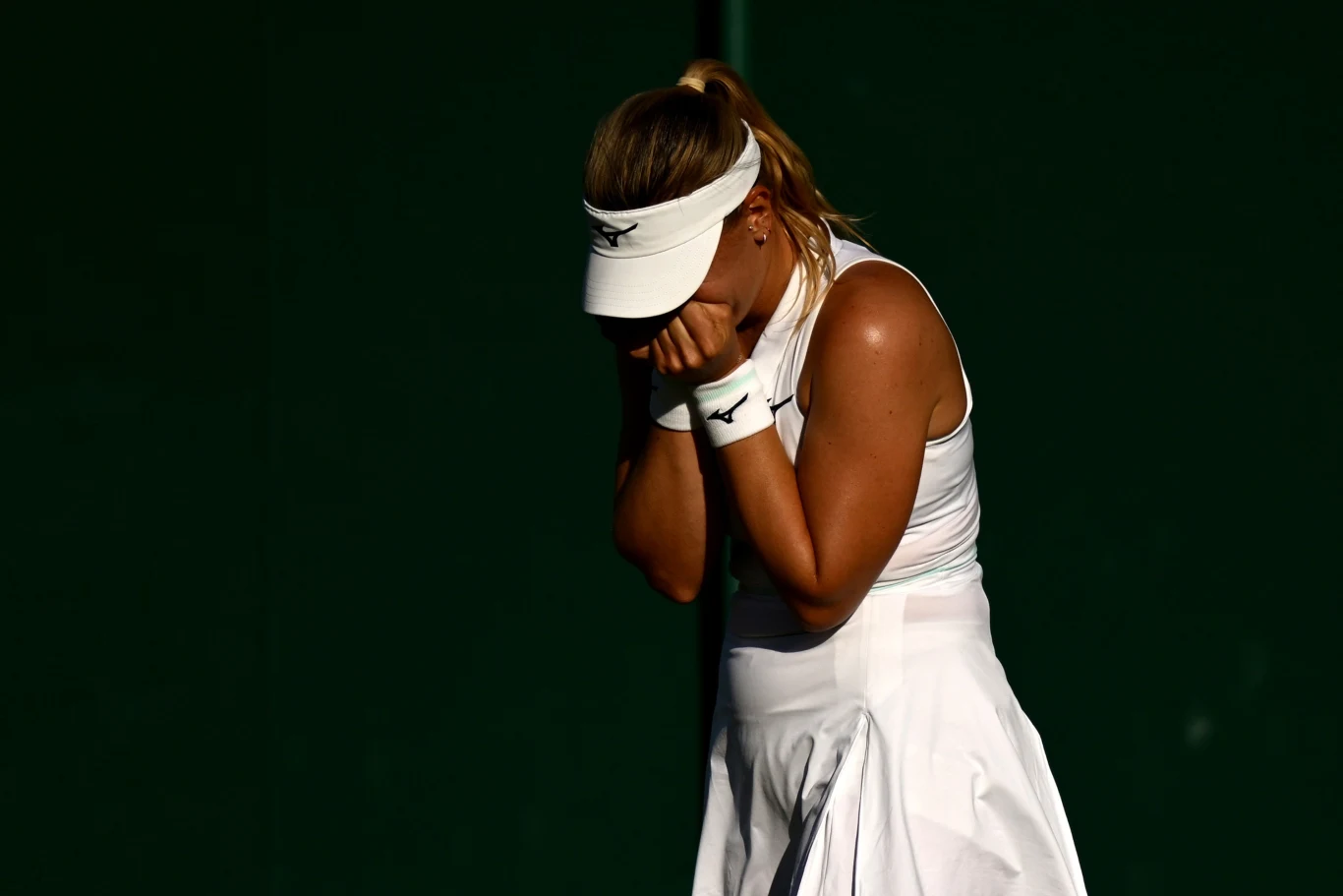 Dalma Galfi rywalizowała z Beatriz Haddad Maią o awans do trzeciej rundy Wimbledonu tenisistka w białej sportowej sukience i czapce zasłania twarz dłońmi w geście rozczarowania na tle ciemnego, jednolitego tła