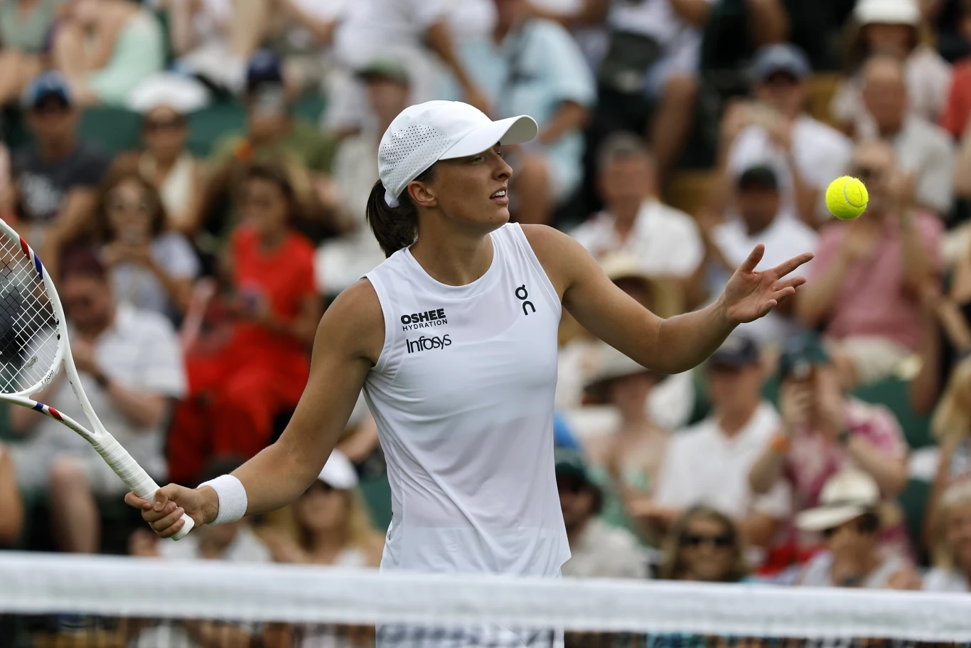 Iga Świątek Tenisistka w białym stroju podczas meczu na korcie, przygotowująca się do odbicia piłki, w tle widoczna publiczność na trybunach.