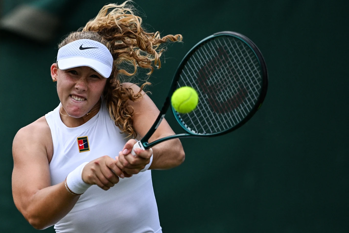 Kobieta wykonuje dynamiczny zamach rakietą tenisową, koncentrując się na zbliżającej się piłce, ubrana w biały strój sportowy i opaskę na głowie, z widocznym logo znanej marki sportowej.