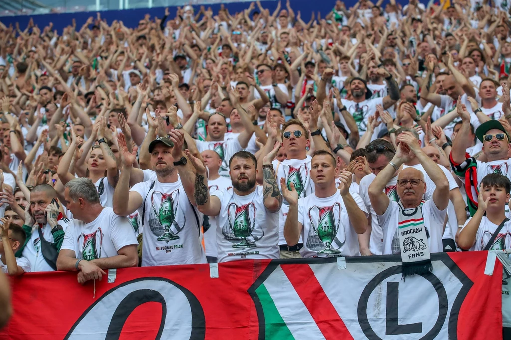 Kibice Legii Warszawa Tłum kibiców piłkarskich ubranych w białe koszulki z barwami i herbem klubu Legia Warszawa, z uniesionymi rękoma, dopingujących swoją drużynę z trybun stadionu, z dużą flagą klubową na pierwszym planie.