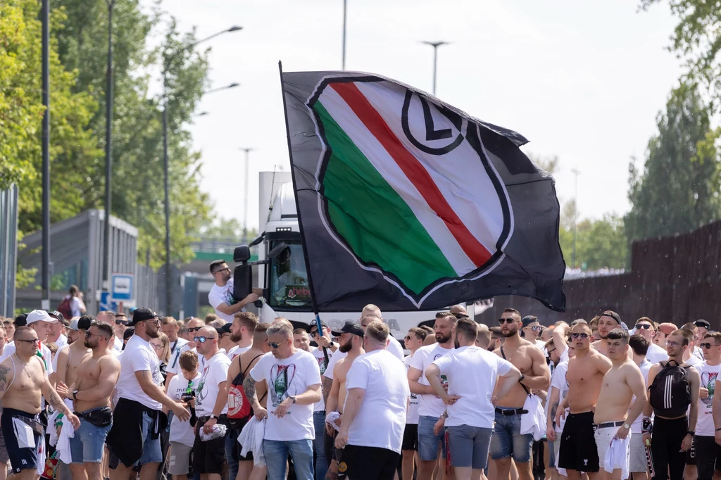 Kibice Legii Warszawa Tłum kibiców maszerujących ulicą, w centrum duża flaga z herbem klubu piłkarskiego Legia Warszawa, większość osób ubrana w jasne koszulki lub bez koszulek, słoneczna pogoda i drzewa w tle.