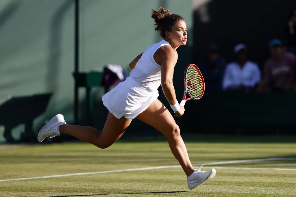 Jasmine Paolini Zawodniczka w białym stroju sportowym biegnie po trawiastym korcie tenisowym, trzymając rakietę w ręce, w tle widoczni są rozmyci kibice.