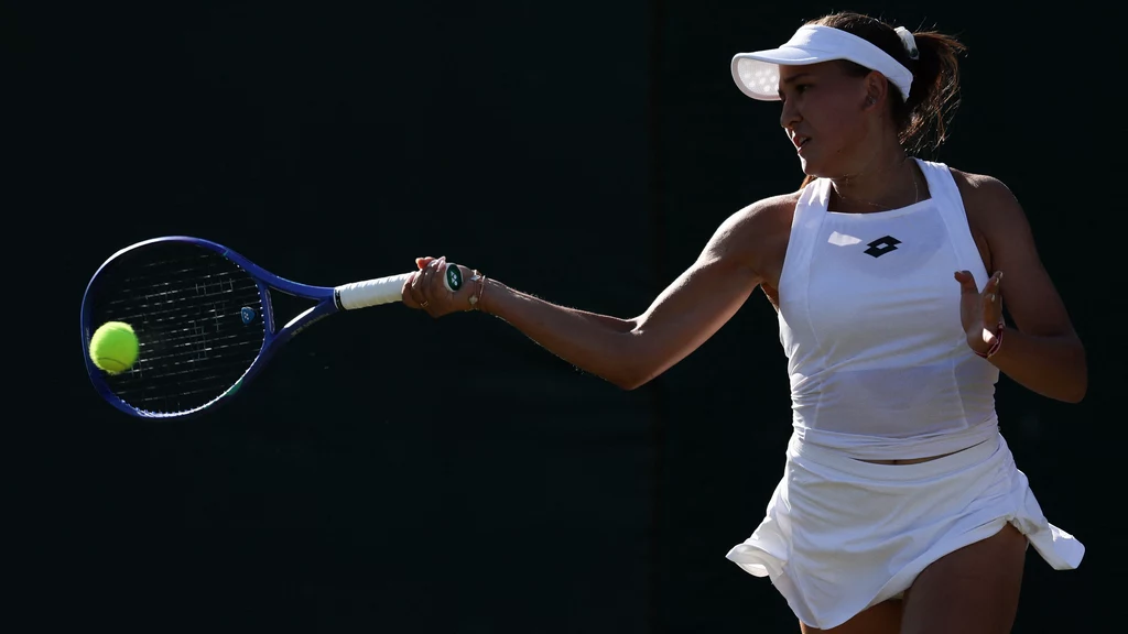 Kamilla Rachimowa Tenisistka ubrana w jasny strój sportowy wykonuje zamach rakietą, przygotowując się do odbicia piłki na korcie tenisowym, światło wyostrza sylwetkę sportowca na ciemnym tle.