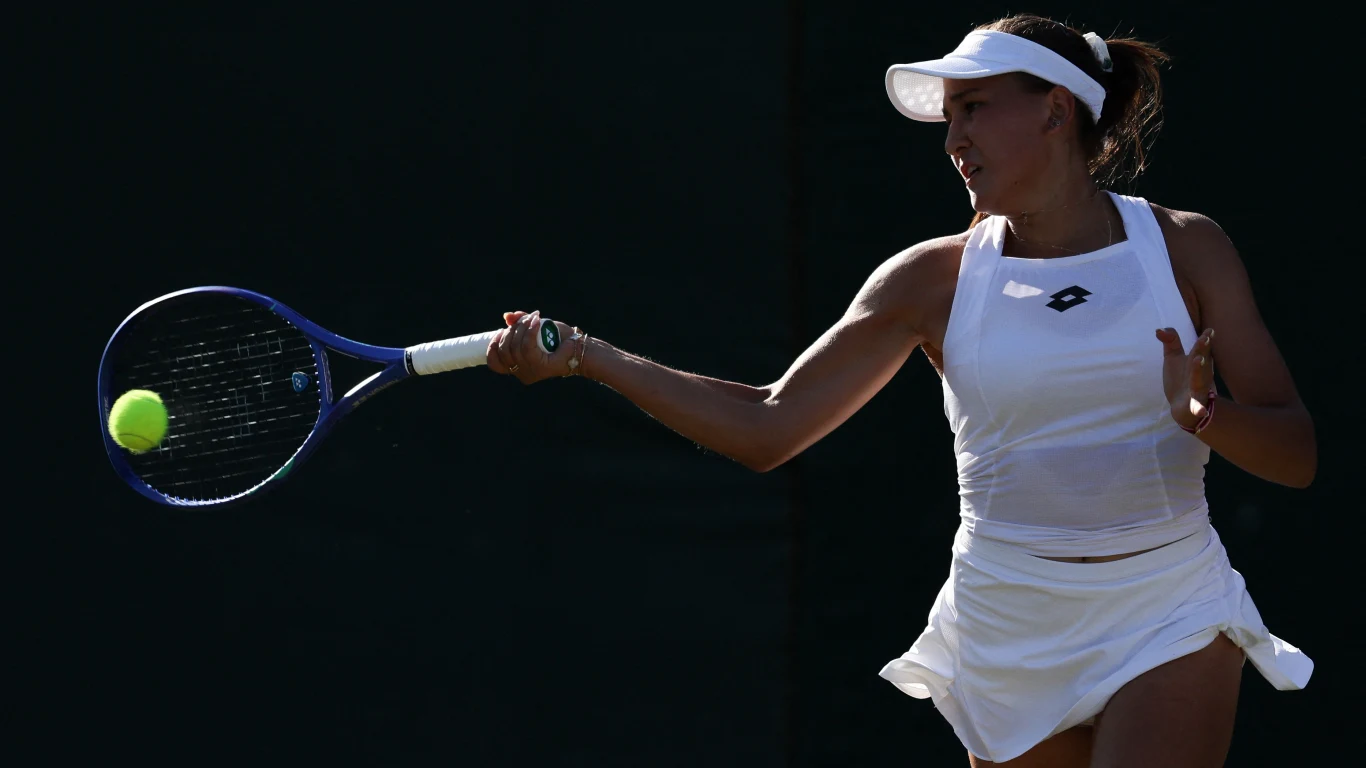 Kamilla Rachimowa Tenisistka ubrana w jasny strój sportowy wykonuje zamach rakietą, przygotowując się do odbicia piłki na korcie tenisowym, światło wyostrza sylwetkę sportowca na ciemnym tle.