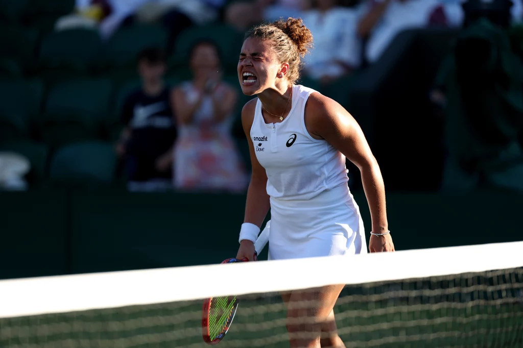 Jasmine Paolini Zawodniczka tenisowa w białej sukience sportowej z rakietą w ręku, wyraża silne emocje na korcie podczas meczu, w tle widownia na stadionie.