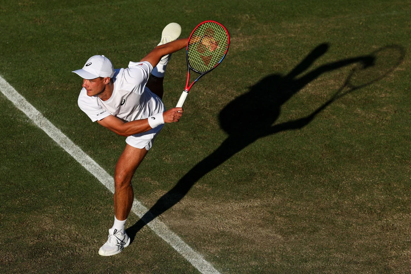 Kamil Majchrzak podczas meczu z Ethanem Quinnem na Wimbledonie, 02.07.2025 r. Tennisista w białym stroju wykonuje serwis na trawiastym korcie, rzucając wyraźny cień na murawę, podkreślający dynamikę ruchu.