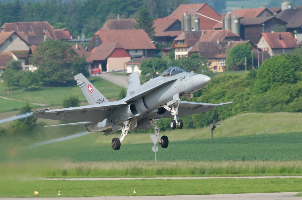 Myśliwiec F/A-18 startujący z bazy lotniczej w Payerne. Siły Powietrzne Szwajcarii posiadają obecnie 30 maszyn tego typu Myśliwiec wojskowy z oznaczeniami Szwajcarii unosi się nad pasem startowym, w tle zielone pola i zabudowania wiejskie o tradycyjnej architekturze.
