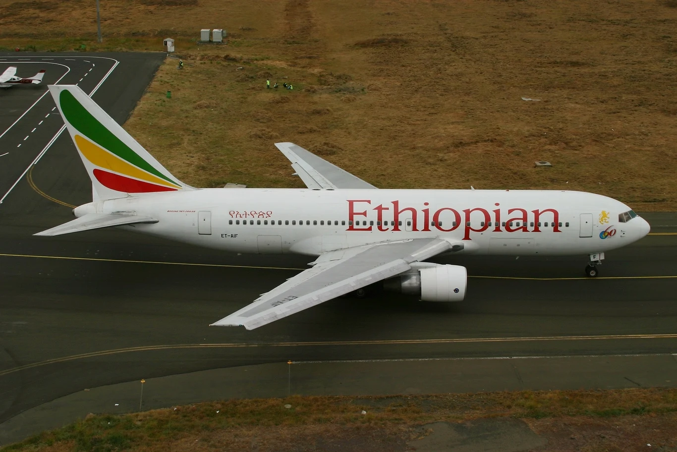 Samolot pasażerski linii Ethiopian Airlines ustawiony na pasie lotniska, charakterystyczne logo oraz kolorowe barwy na ogonie, otoczenie stanowi płyta lotniska i pas startowy.