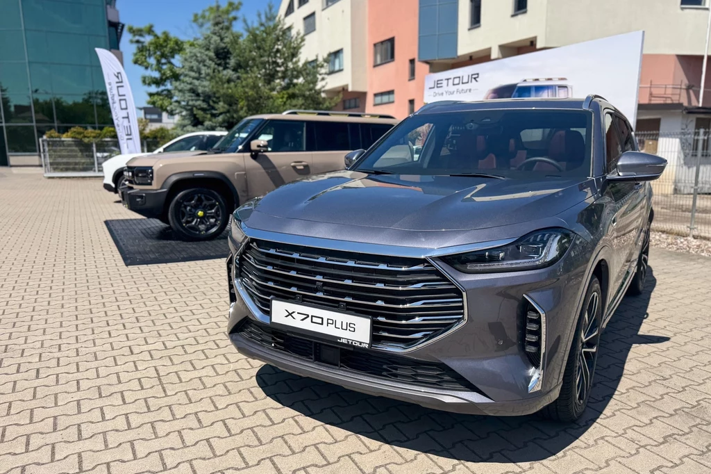 Dwa nowoczesne samochody marki Jetour zaparkowane na placu przed budynkiem, na pierwszym planie szary SUV X70 Plus, w tle drugi model pojazdu oraz banery reklamowe.