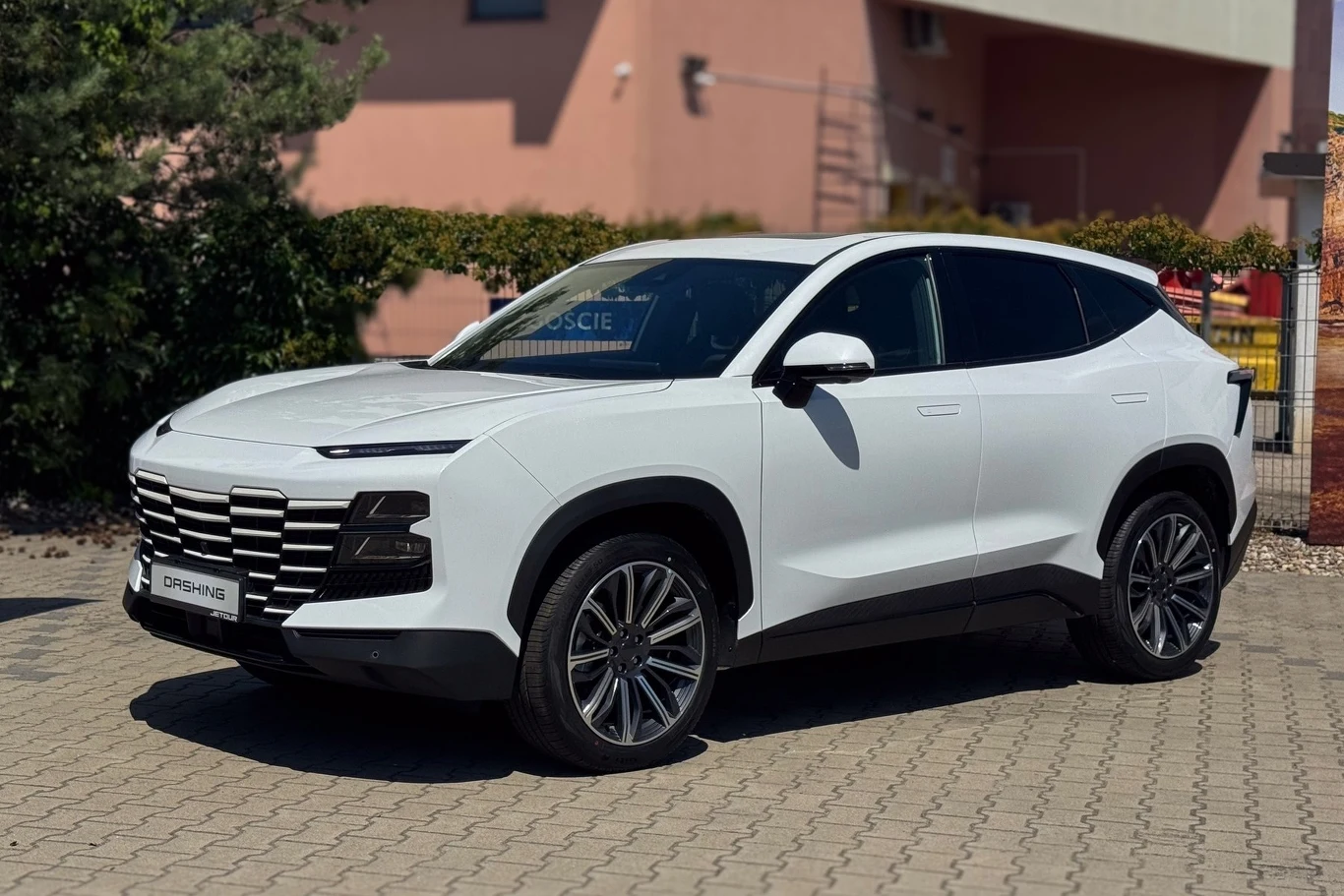 Jetour Dashing bryłą i detalami może przypominać trochę Lamborghini Urus. Chiński SUV prezentuje się bardzo nowocześnie. Biały SUV Jetour Dashing zaparkowany na chodniku, w tle fragment budynku i roślinność, charakterystyczne ostre linie karoserii oraz nowoczesny design pojazdu.