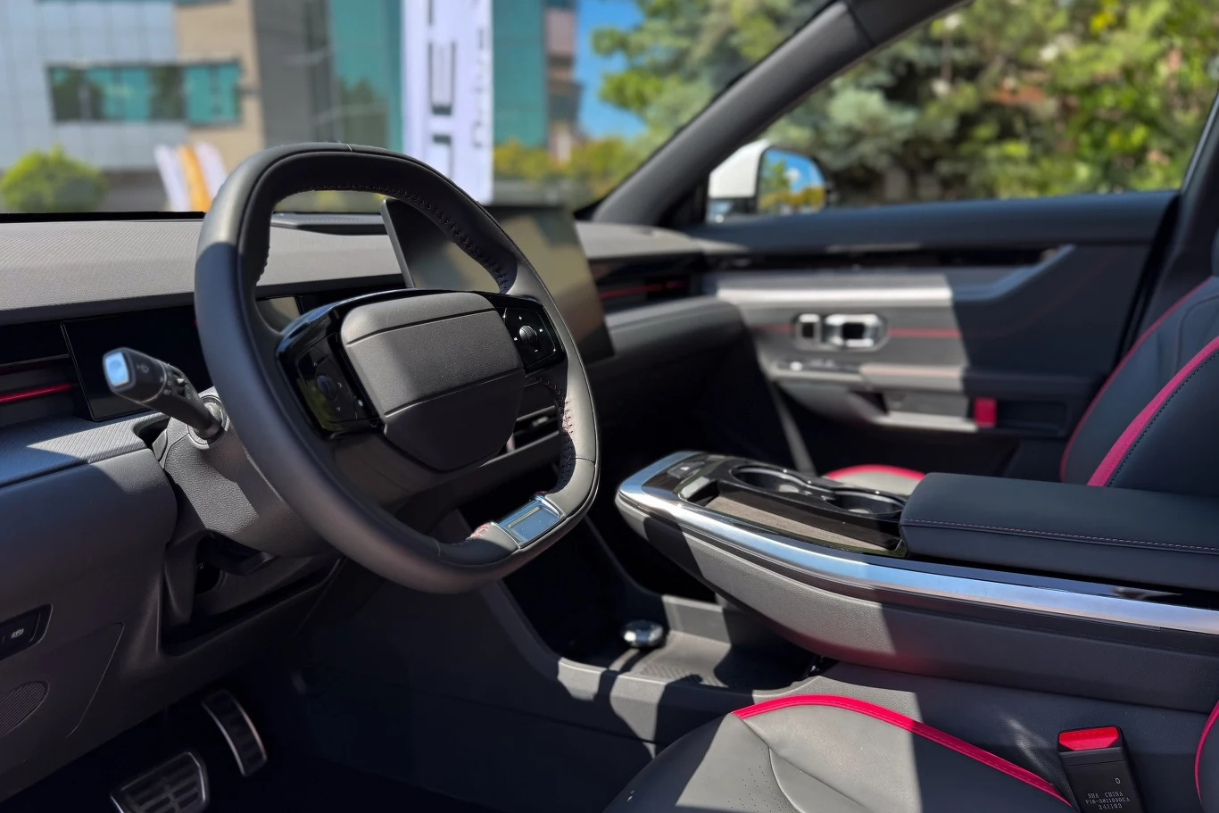 Nowoczesna kabina samochodu z czarną kierownicą, designerską deską rozdzielczą, ekranem dotykowym oraz komfortowymi fotelami z czerwonymi przeszyciami, w tle widać budynki i zieleń za szybą.