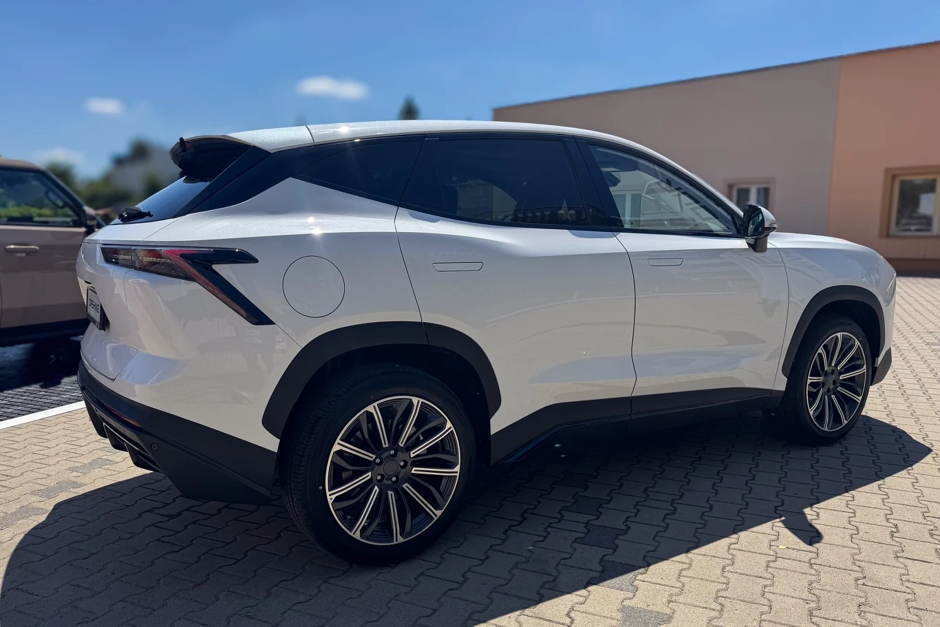 Biały nowoczesny SUV zaparkowany na utwardzonym placu, samochód stoi przy jasnym budynku, w tle widoczne są fragmenty innych aut oraz niebo z chmurkami.