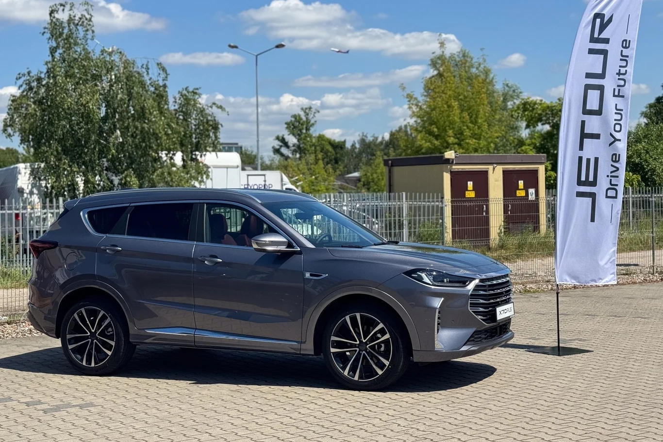Jetour X70 Plus Srebrny samochód osobowy SUV marki Jetour stoi na parkingu na tle zieleni i elementów infrastruktury miejskiej, obok powiewająca wysoka flaga z logo producenta.