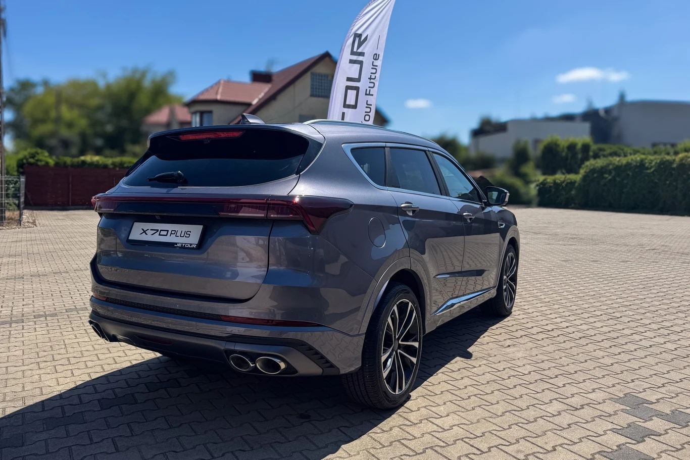 Szary samochód marki X70 Plus typu SUV zaparkowany na brukowanym placu na tle niskiej zabudowy i zieleni, z widoczną flagą reklamową obok pojazdu.