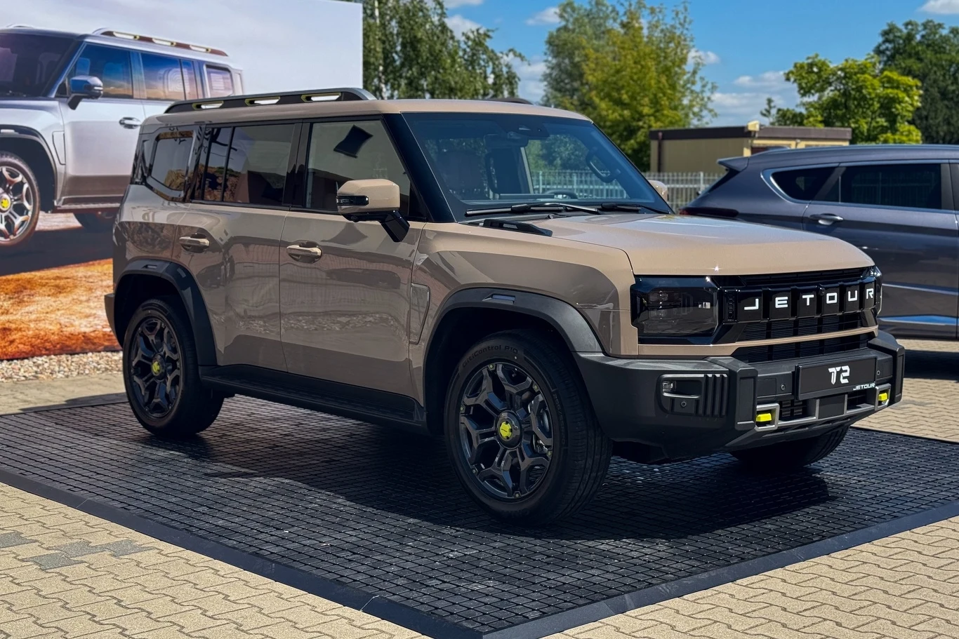 Jetour T2 Nowoczesny SUV w kolorze beżowym marki Jetour T2, ustawiony na ekspozycji plenerowej, z dużymi felgami i przyciemnianymi szybami, w tle tablica reklamowa z tym samym modelem oraz fragment innego pojazdu.