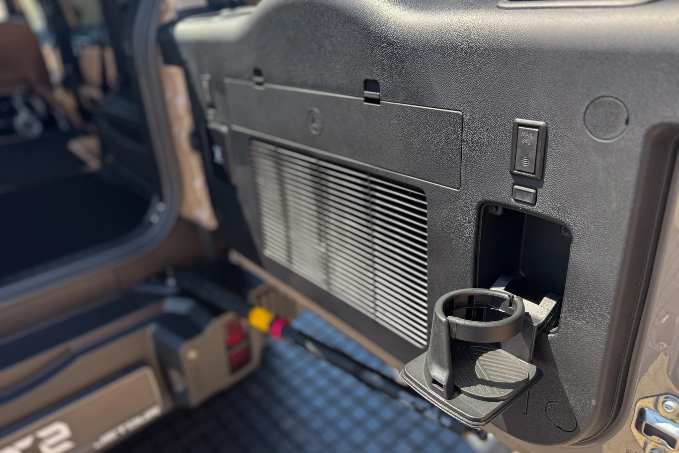 Wnętrze tylnej części pojazdu z wyraźnie widocznym uchwytem na napoje zamontowanym na drzwiach oraz kratką wentylacyjną, w tle fragment siedzenia i podłogi bagażnika.