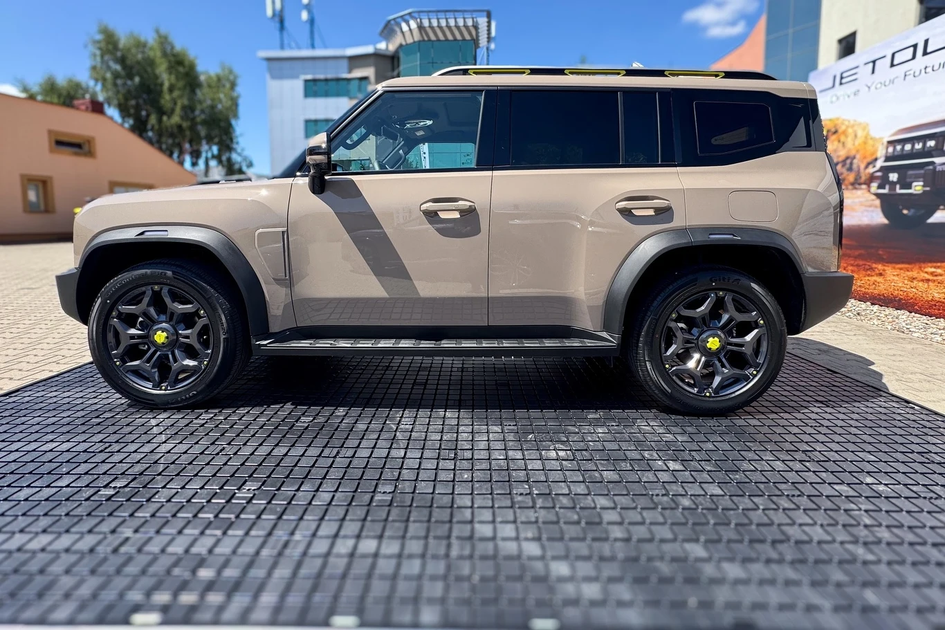 Beżowy samochód terenowy typu SUV, stojący na czarnej macie ekspozycyjnej na tle zabudowań i plakatu reklamowego, z dużymi czarnymi felgami i przyciemnianymi szybami.