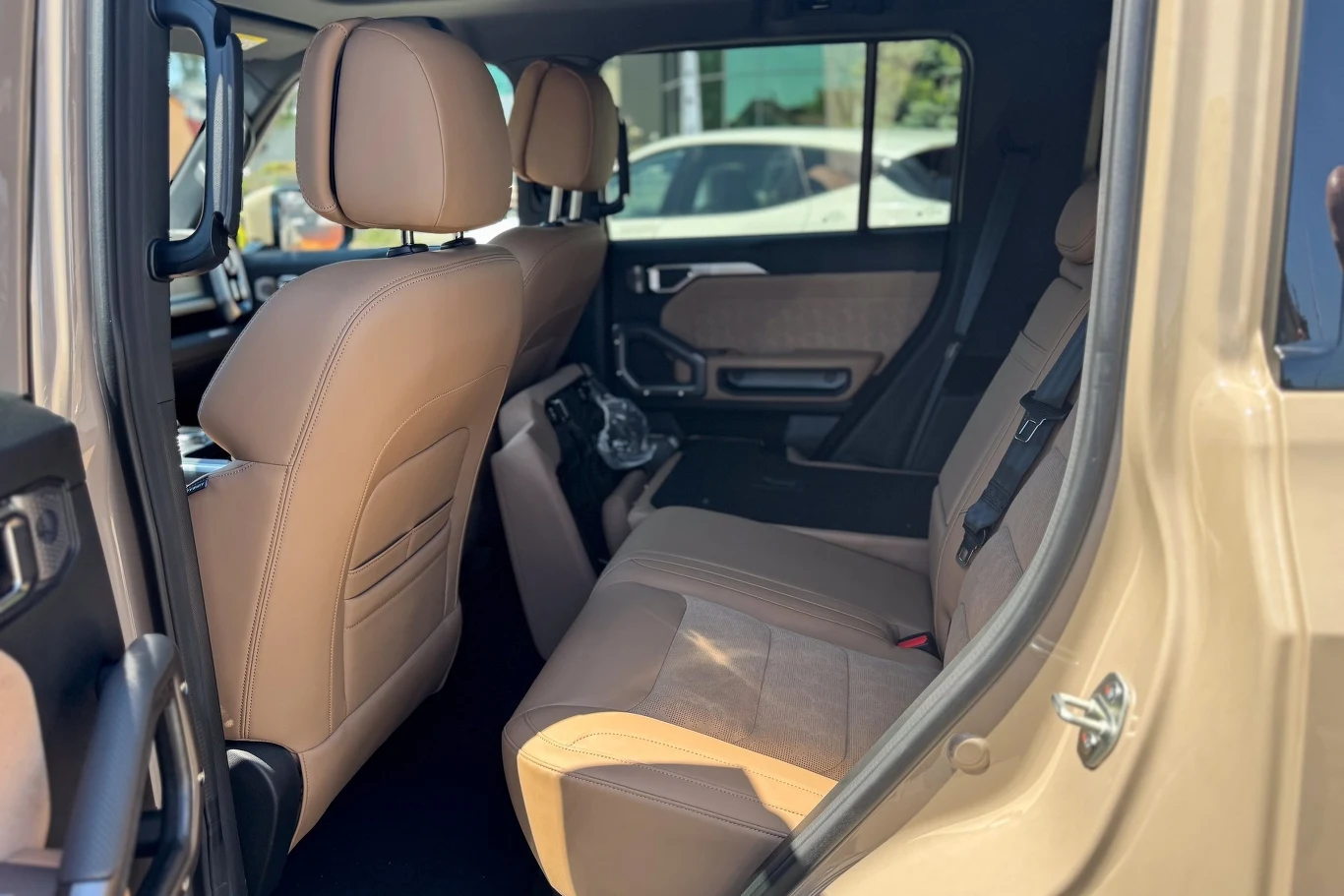 Wnętrze samochodu z beżową tapicerką, widok na tylne siedzenia i przednie fotele, otwarte drzwi tylne, w tle przez okno widoczne fragmenty innych pojazdów.