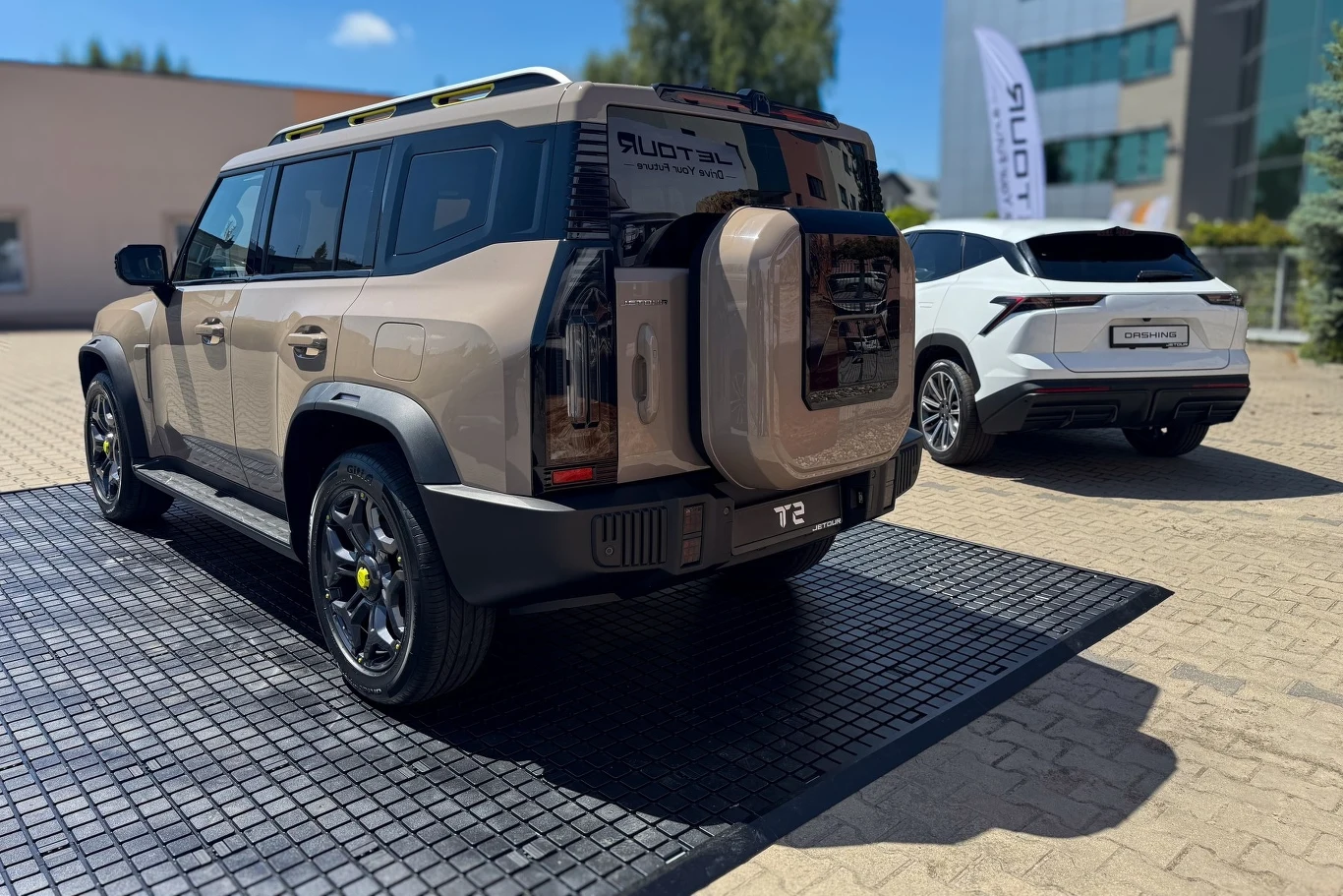 Dwa nowoczesne samochody terenowe zaparkowane obok siebie na podłożu wyłożonym matą, w tle widoczne budynki i elementy miejskiego otoczenia, jasny dzień i dobrze widoczne detale karoserii pojazdów.