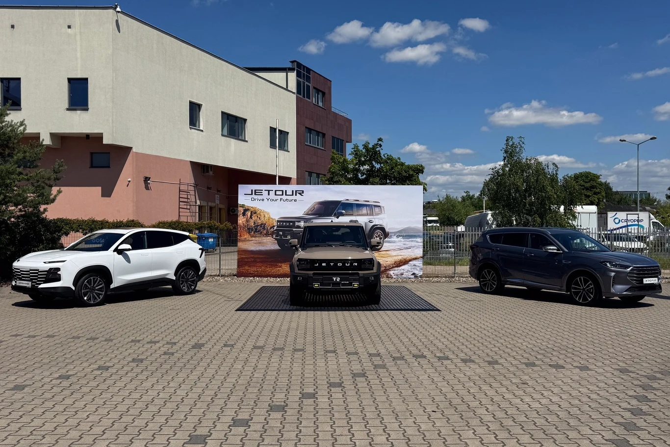 Trzy samochody marki Jetour ustawione na placu przed banerem prezentującym logo firmy oraz zdjęcie jednego z modeli, po bokach stoją białe i ciemne SUV-y, w centrum ciemny model z wyróżniającym się wyrazistym grillem, w tle budynek przemysłowy oraz zie...
