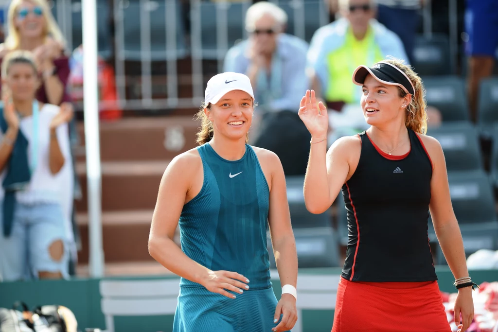 Iga Świątek i Caty McNally wspólnie wygrały juniorski turniej French Open - w deblu Dwie kobiety ubrane w sportowe stroje tenisowe znajdują się na korcie, obie są uśmiechnięte, jedna z nich unosi rękę w geście powitania lub pożegnania, w tle widoczni są kibice na trybunach.