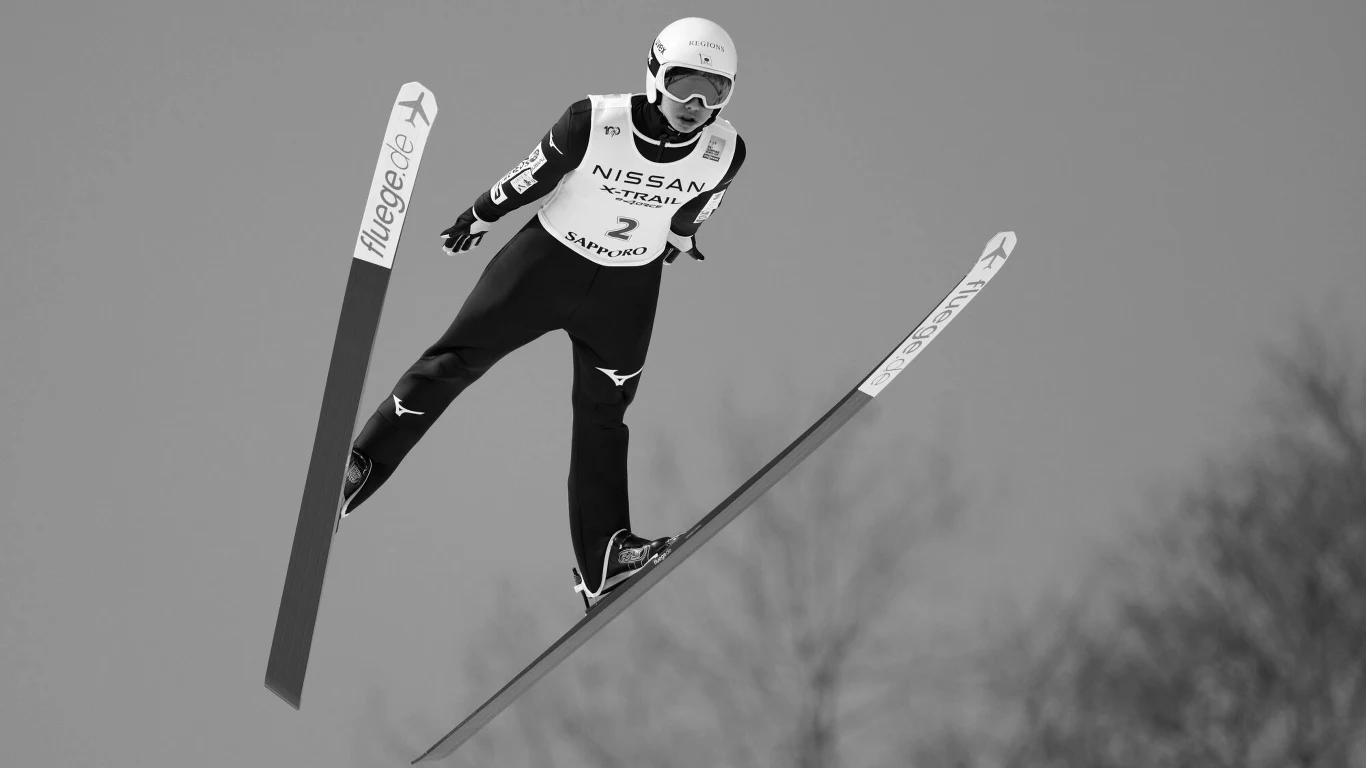 Nie żyje Asahi Sakano Skoczek narciarski w powietrzu podczas skoku z dużą prędkością, w specjalnym kombinezonie, z kaskiem i goglami na głowie, wyraźnie oznaczone narty, rozmazane tło wskazujące na dużą wysokość i dynamikę ruchu.