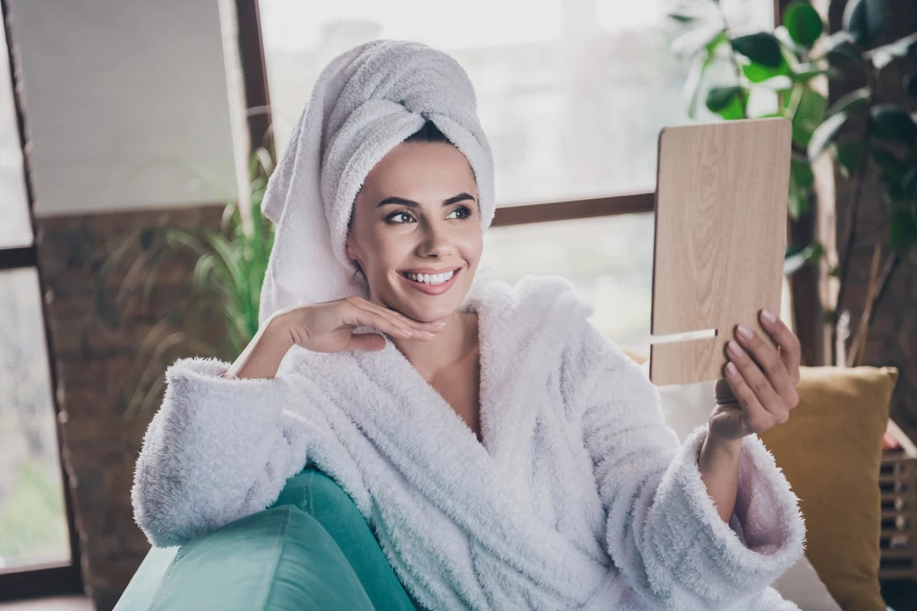 Włoskie spa? Wystarczą trzy produkty, żeby dobrze wyglądać Uśmiechnięta kobieta w białym szlafroku i turbanie na głowie siedzi na sofie, trzymając w ręce lustro i przyglądając się swojemu odbiciu; w tle rośliny doniczkowe i duże okna.