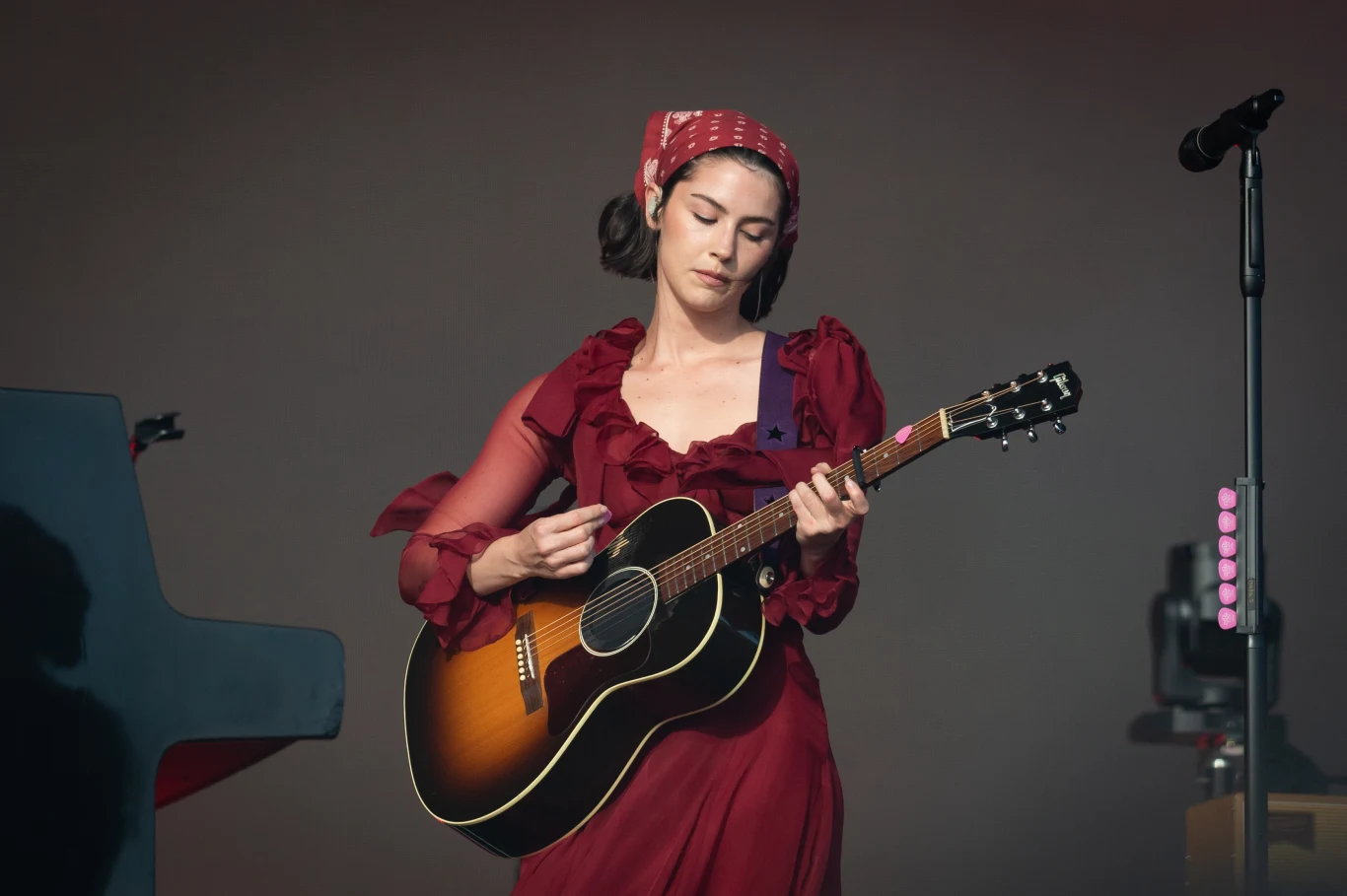 Gracie Abrams pierwszy raz wystąpi w Polsce na Open'er Festivalu 2025 Kobieta w czerwonej sukience i opasce na głowie gra na gitarze akustycznej na scenie, obok mikrofon na statywie oraz instrument klawiszowy po lewej stronie kadru