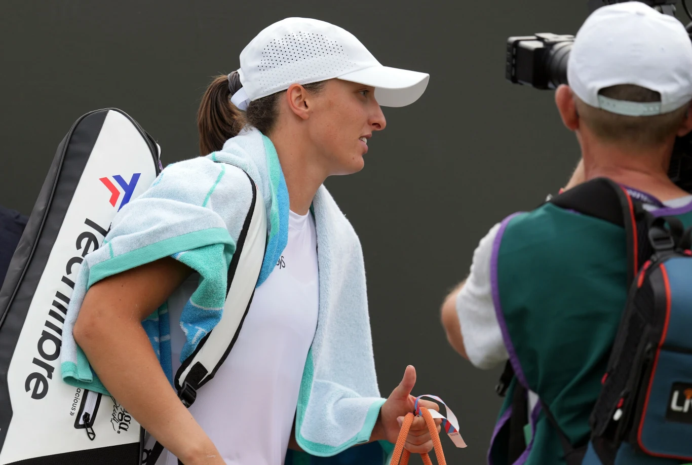 Iga Świątek Tenisistka ubrana w sportowy strój i białą czapkę z daszkiem przechodzi obok fotografa, na ramieniu niesie torbę z rakietą i ręcznik, w tle neutralna powierzchnia kortu.