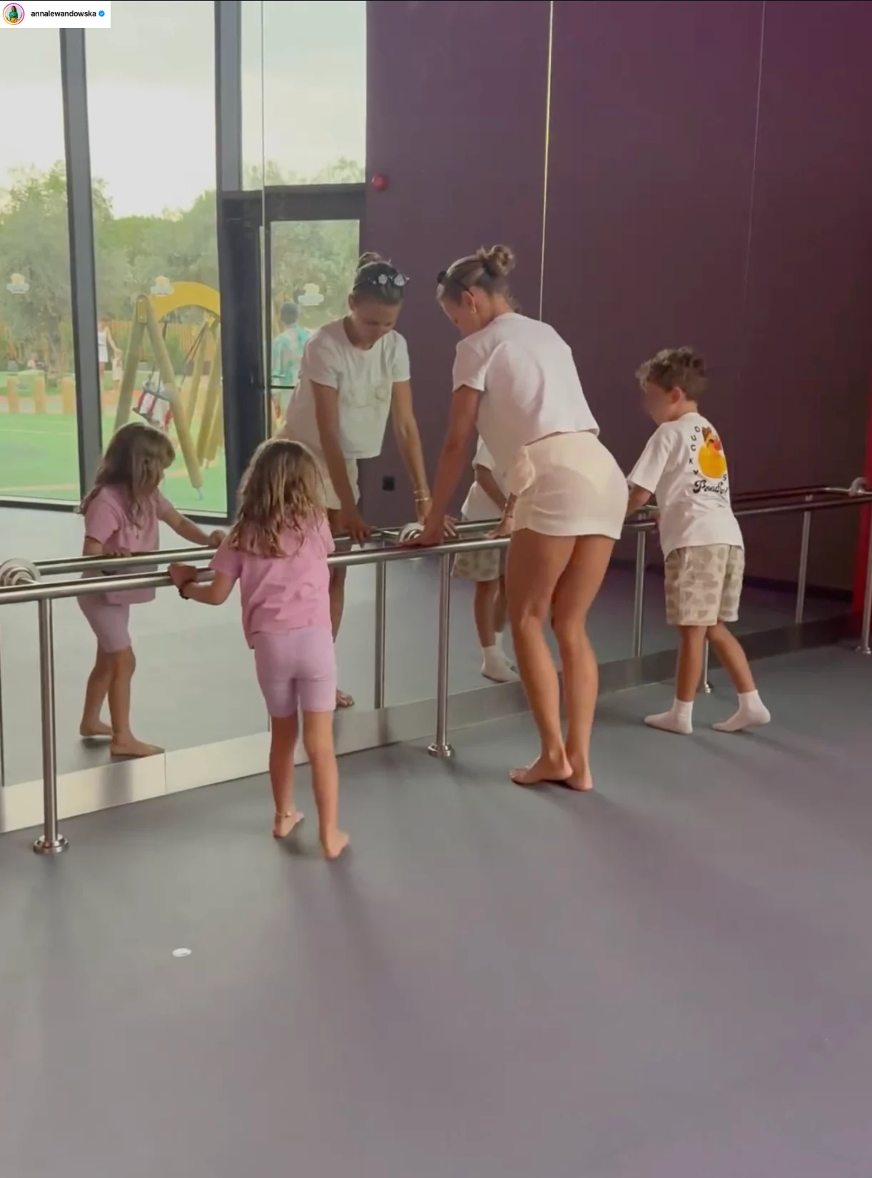 Anna Lewandowska z córką Laurą i Liamem Szczęsnym podczas treningu Kobieta oraz dwoje dzieci ćwiczą przy drążku baletowym ustawionym przy dużym lustrze, w tle widoczne okno z widokiem na plac zabaw na zewnątrz, ściany sali mają fioletowy kolor.