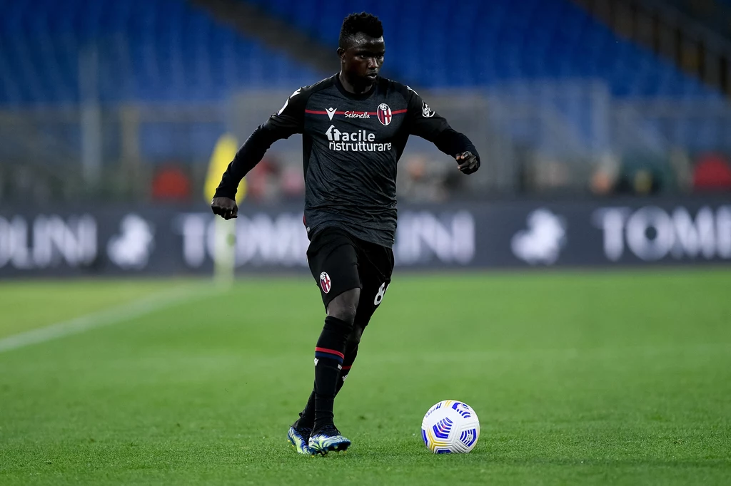 Musa Juwara w barwach Bologna FC podczas meczu Serie A z AS Roma Piłkarz w czarnej koszulce drużyny Bologna FC biegnie po murawie stadionu podczas meczu, kontrolując piłkę przy nodze. W tle widoczne rozmyte trybuny i fragmenty reklam przy boisku.