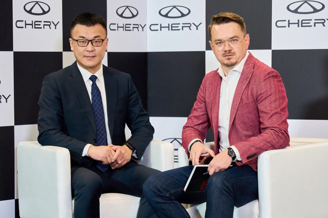 Dwóch mężczyzn w formalnych ubraniach siedzi w białych fotelach na tle firmowych logotypów Chery. Jeden trzyma notes, obaj patrzą w stronę obiektywu.
