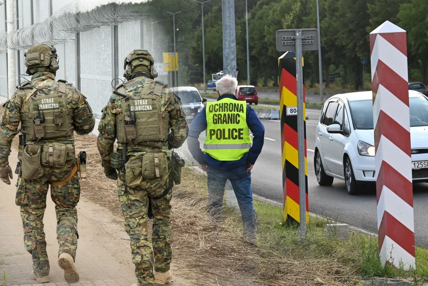 Polska przywraca kontrole na granicach z Niemcami i Litwą (Zdj. ilustracyjne) Dwóch funkcjonariuszy Straży Granicznej w kamuflażu patroluje teren przy ogrodzeniu granicznym, w tle osoba w kamizelce z napisem 'Ochrona Granic Police' stoi przy słupku granicznym oraz przejeżdżających samochodach.