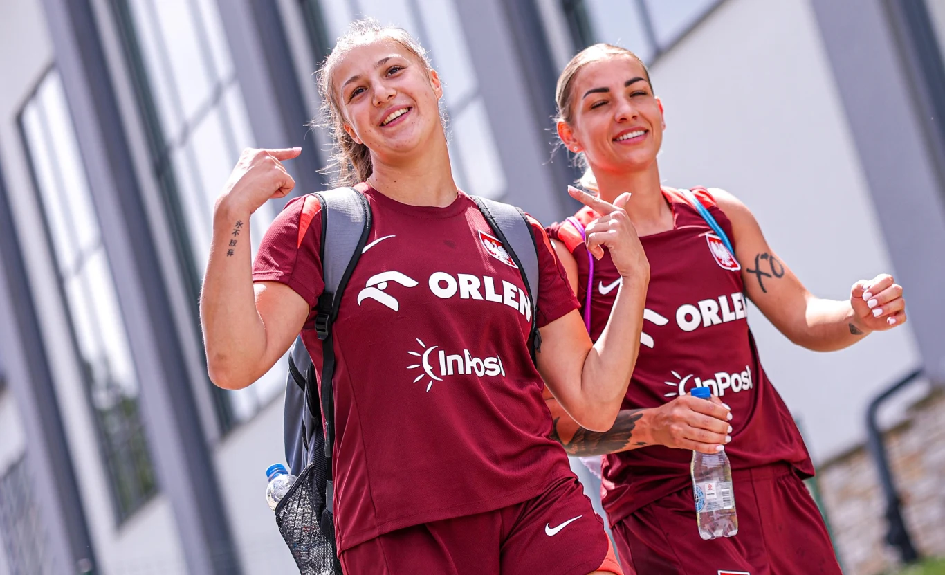 Emilia Szymczak (z lewej) Dwie uśmiechnięte piłkarki w bordowych strojach sportowych z logiem Orlen i InPost, niosące sportowe plecaki, przechadzające się w pobliżu nowoczesnego budynku, jedna trzyma butelkę wody.