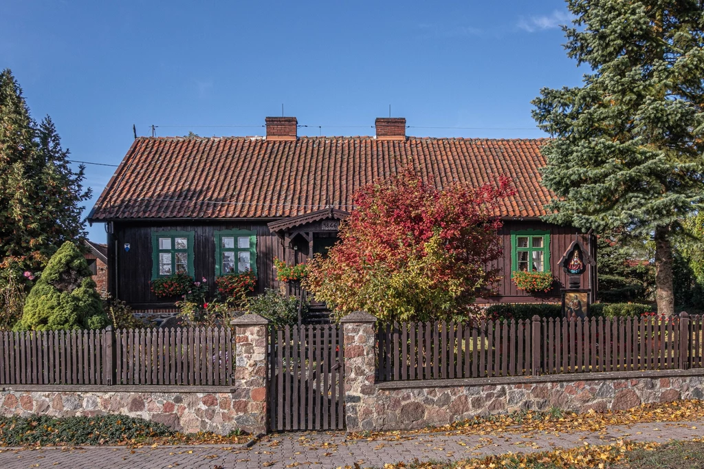 Drewniany dom z czerwonym dachem otoczony ogrodem z kolorowymi krzewami i drzewami, całość ogrodzona kamienno-drewnianym płotem, pod błękitnym niebem.