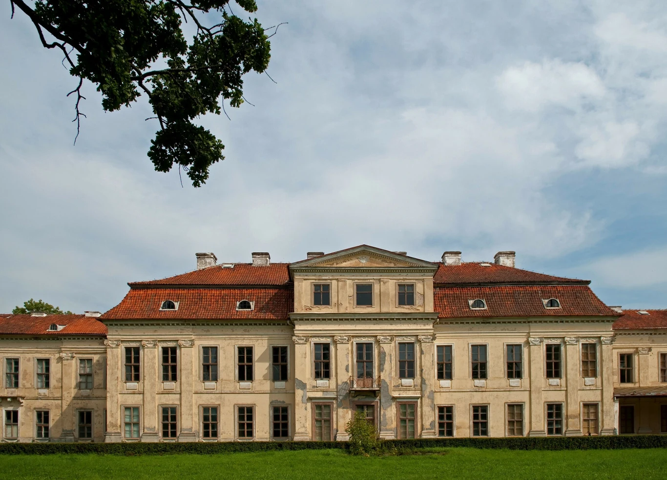 Barokowy pałac z jasną elewacją i czerwonym dachem położony w otoczeniu zieleni, z szerokim trawnikiem na pierwszym planie, na tle nieba z delikatnymi chmurami.