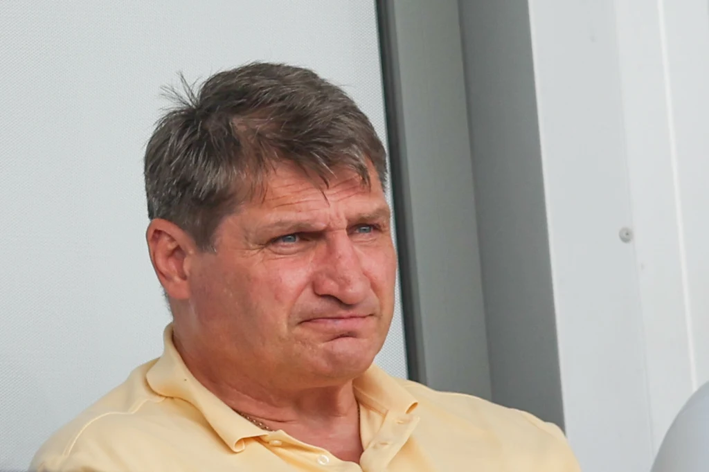 Andrzej Gołota Mężczyzna w średnim wieku w jasnej koszuli siedzi i patrzy przed siebie z poważnym wyrazem twarzy, tło stanowi jasna ściana.