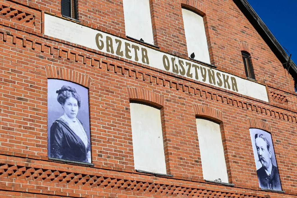 Ceglana fasada budynku z dużym szyldem Gazeta Olsztyńska oraz dwoma czarno-białymi portretami historycznych postaci umieszczonymi po obu stronach elewacji.