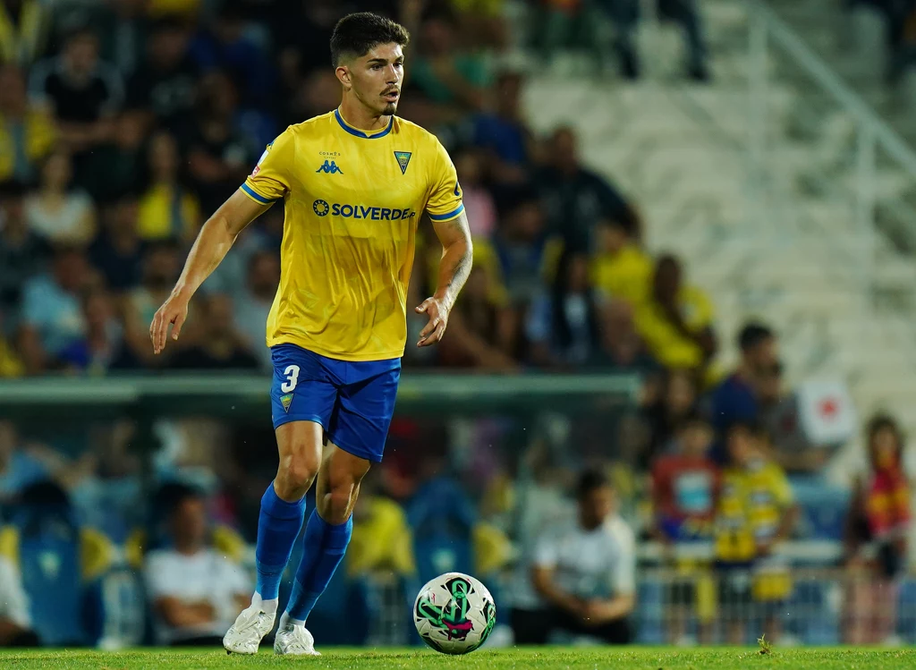 Bernardo Vital w barwach Estoril Piłkarz w żółtej koszulce i niebieskich spodenkach kontroluje piłkę na boisku podczas meczu, wokół niego widoczni są kibice na trybunach.