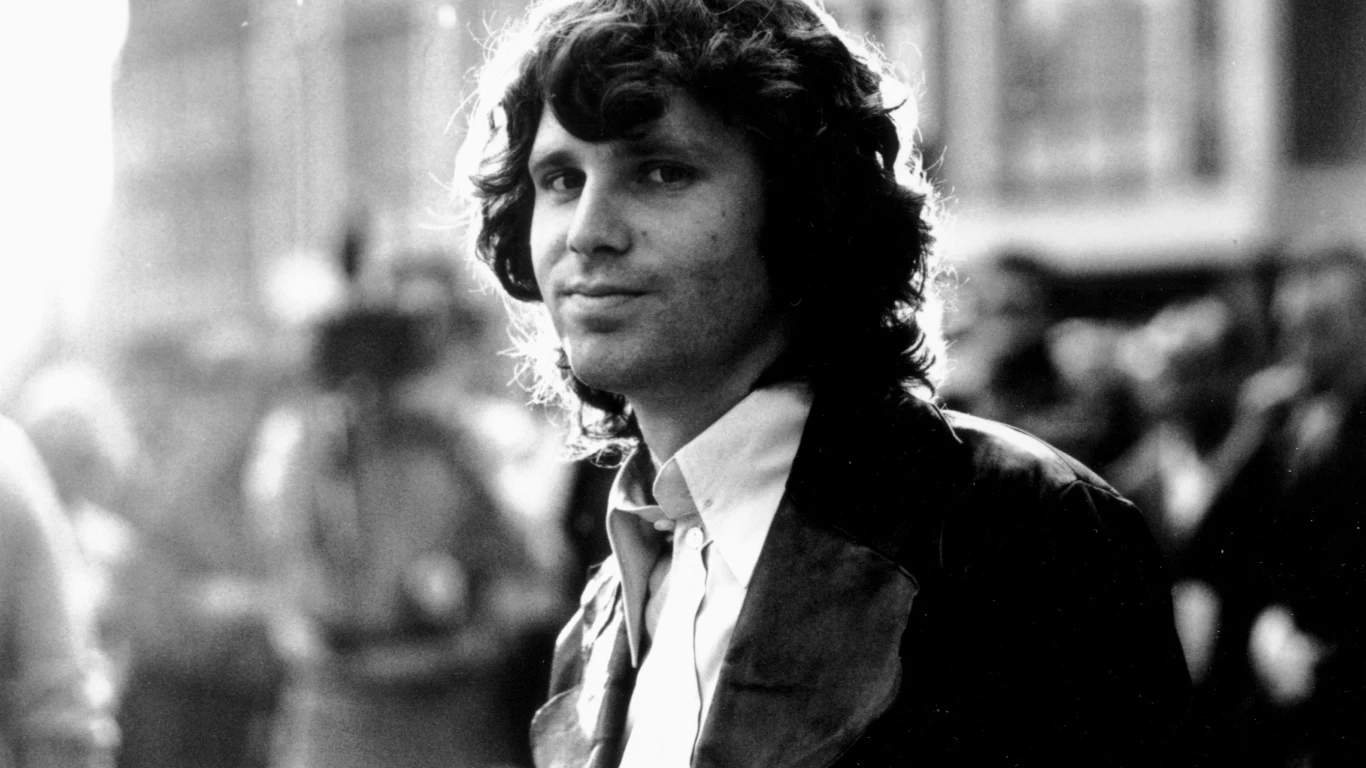 Jim Morrison (1968) Mężczyzna z długimi, kręconymi włosami ubrany w biały koszulę i ciemną kurtkę patrzy przed siebie, w tle niewyraźna sylwetka ludzi oraz rozmyte miejskie otoczenie.
