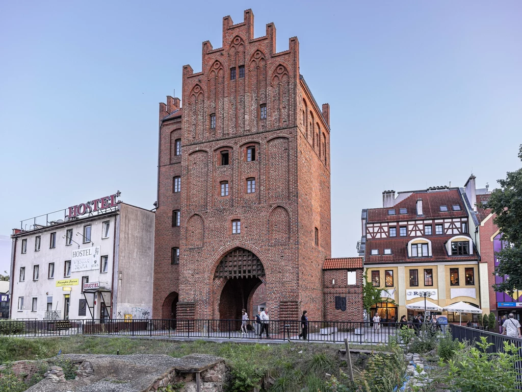 Gotycka brama miejska z czerwonej cegły z ostrołukowym wejściem, otoczona niższymi budynkami mieszkalnymi i hostelem, w tle jasne niebo, a przed bramą przechodzący ludzie